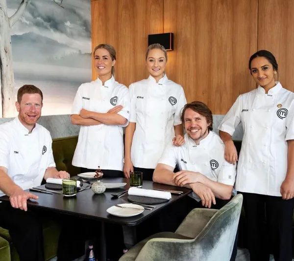 MasterChef finalists in white chef coats smiling, standing together in a dining area, with one seated at a table.