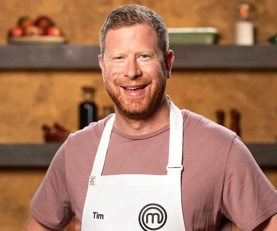 Man in a MasterChef apron smiling in a kitchen setting.