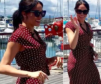 A woman in a polka dot dress and sunglasses smiles and waves on a pier with boats in the background.