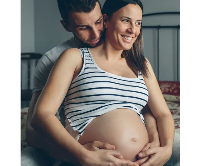 Pregnant woman in striped tank top, smiling, embraced by partner from behind, hands on belly.