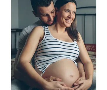 Pregnant woman in striped tank top, smiling, embraced by partner from behind, hands on belly.