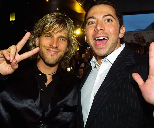 Two people smiling at a public event, one showing a peace sign, wearing suits in a lively atmosphere.