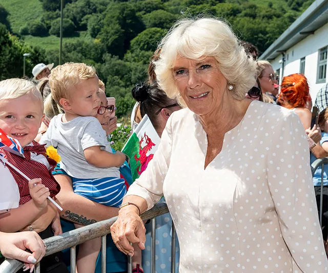 Camilla in a white polka dot dress greeting children and a crowd outdoors on a sunny day.