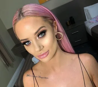 Woman with long pink hair and hoop earrings, wearing makeup, smiles while posing indoors.