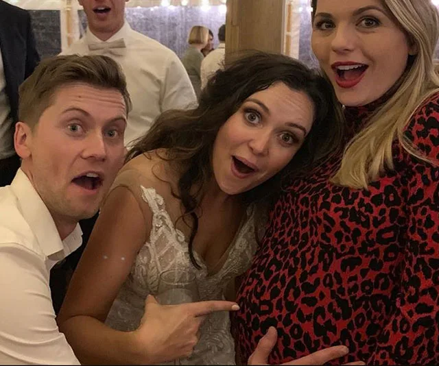 Three people at a celebration: a man and two women, one wearing a red dress and holding her pregnant belly.