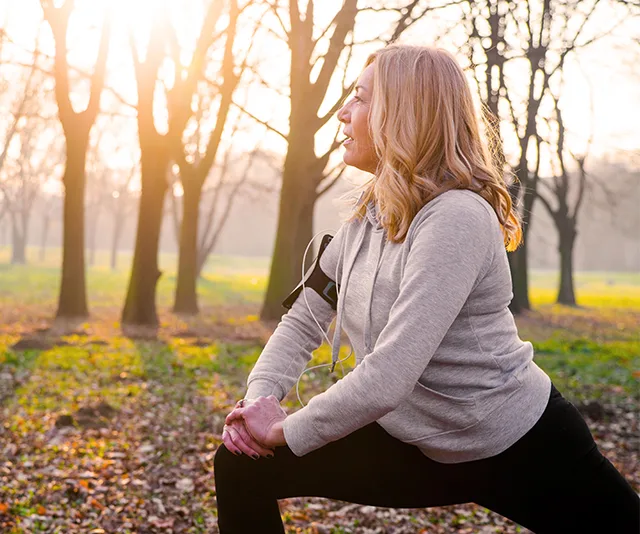 Middle-aged woman stretching in a sunlit park, wearing a hoodie, preparing for a winter workout.