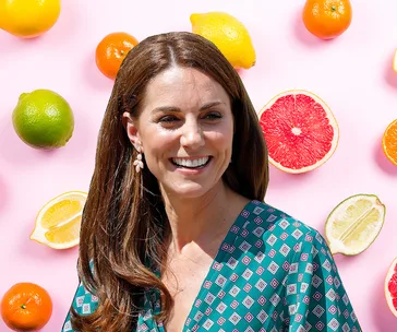 A woman smiling in front of a pink backdrop with colorful citrus fruits scattered around.