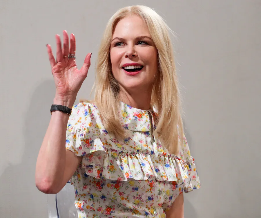 A woman with blonde hair, wearing a floral top, smiles and waves her hand.