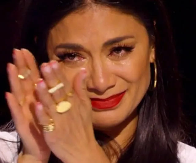 A person with long hair clapping tearfully, wearing red lipstick and gold rings, possibly reacting emotionally.