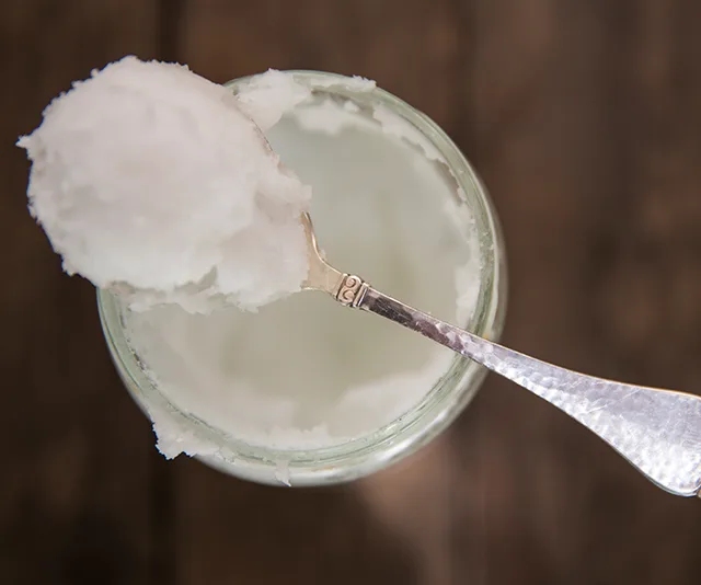 Spoonful of coconut oil over an open jar on a wooden surface.