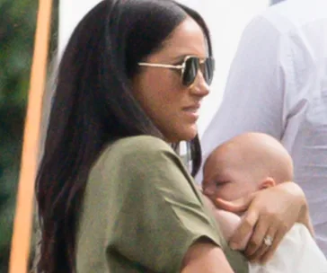 A woman with dark hair and sunglasses holds a baby gently in her arms outdoors.