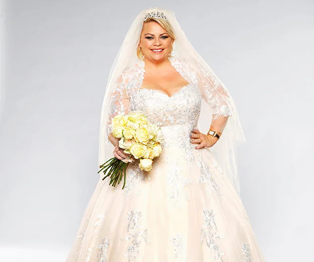 A bride in an ornate white wedding dress holds a bouquet of yellow roses, smiling against a plain background.
