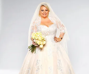 A bride in an ornate white wedding dress holds a bouquet of yellow roses, smiling against a plain background.