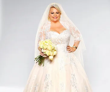A bride in an ornate white wedding dress holds a bouquet of yellow roses, smiling against a plain background.
