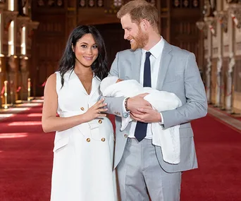 Man and woman with a baby in a hall, woman in white dress, man in grey suit, smiling, red carpet background.