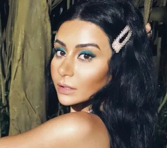 A woman with long dark hair and blue eye makeup, wearing a pearl hair clip, poses outdoors.