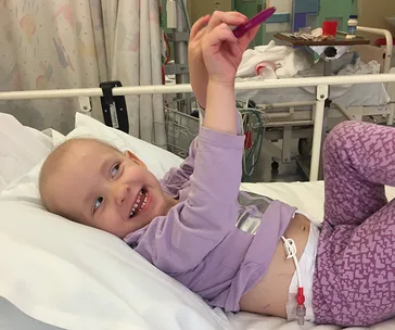 A child smiling and holding a toy in a hospital bed, wearing a purple outfit.