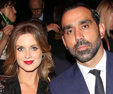 Two people pose together at an event, one wearing a suit and tie, the other with red lipstick and a black dress.