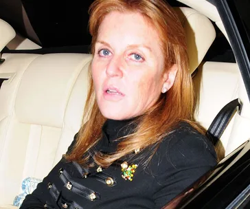 Red-haired woman sitting in a car, wearing a dark top with decorative accents, looking towards the camera.