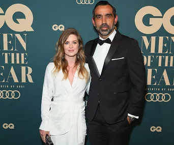 Man in tuxedo and woman in white dress at a GQ Men of the Year event backdrop.