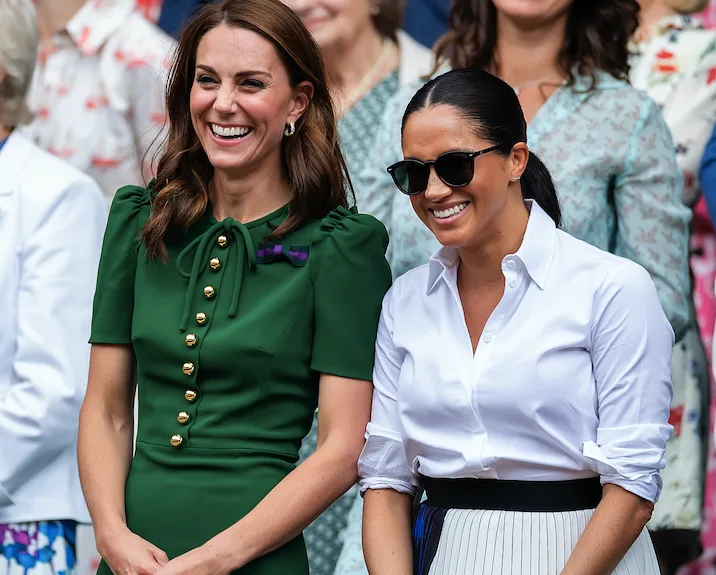 Two women smiling, one in a green dress and the other in a white shirt and sunglasses, standing in a crowd.