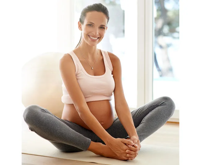 Smiling pregnant woman in workout clothes sitting cross-legged on the floor indoors.