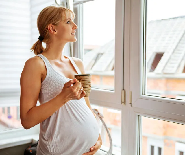Pregnant woman at 25 weeks holding a mug, looking out of a window with a peaceful expression.