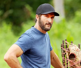 Man in a blue shirt and cap outdoors, near a crafted wooden structure, with greenery in the background.