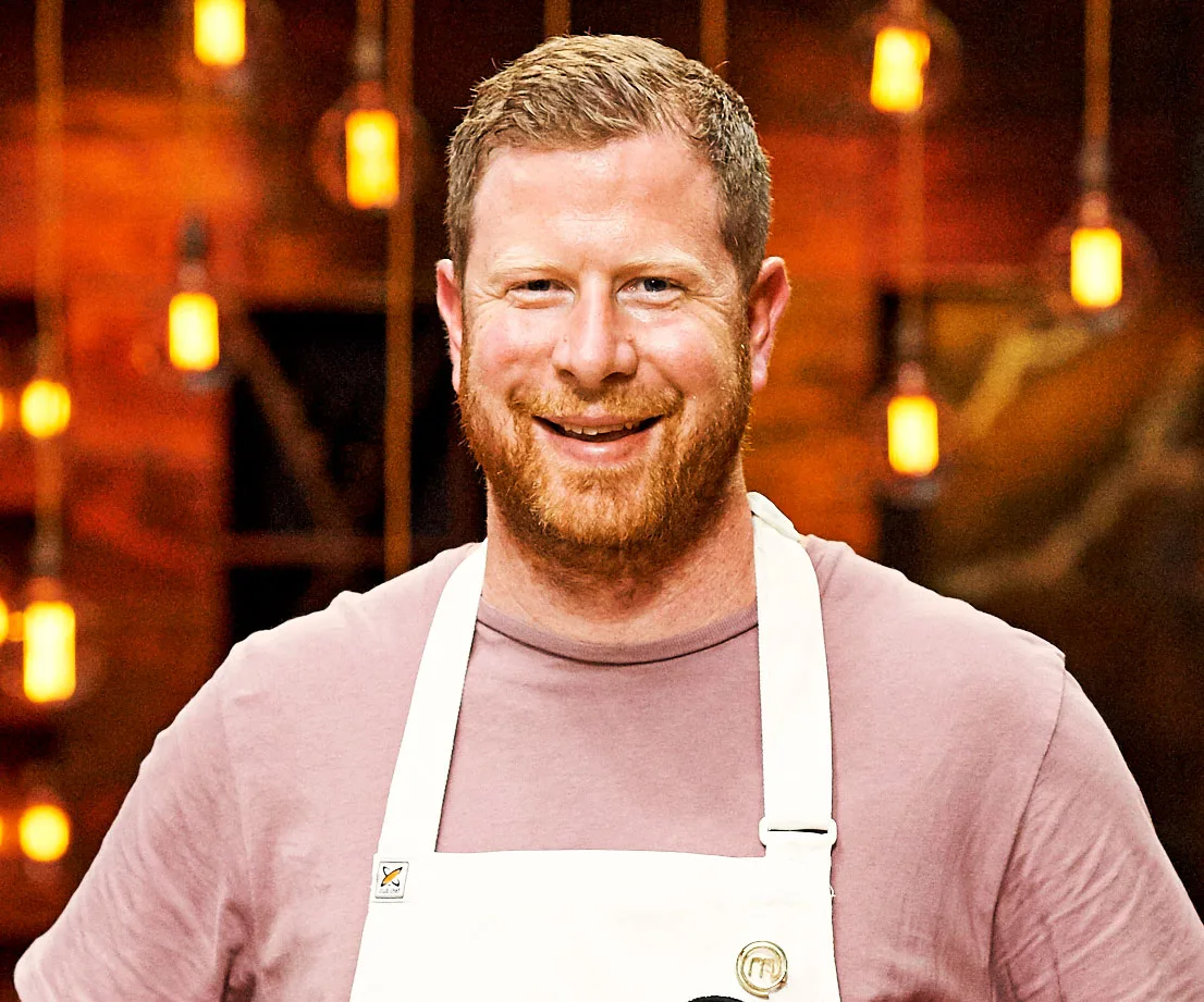 Man with a beard smiling, wearing a white apron, standing in a warmly lit room.