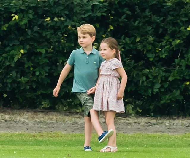 Two young children walking together on a grassy area, with greenery in the background.