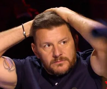 A man with short hair and a beard, holding his head with both hands, looking pensive.