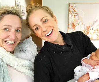 Two women smiling, each holding a baby in their arms, standing indoors with a colorful painting on the wall behind them.