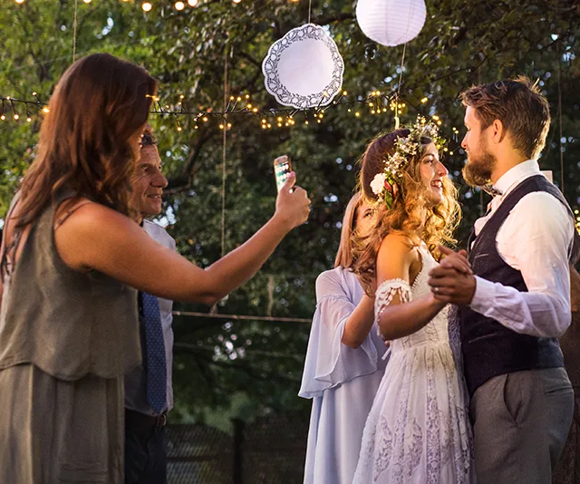 A bride and groom dance at an outdoor wedding, surrounded by guests taking photos under string lights and paper lanterns.
