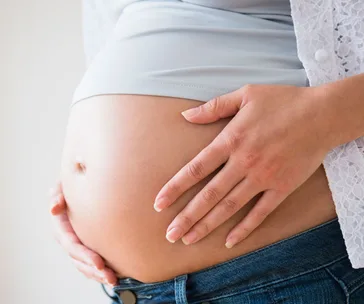 Pregnant woman at 26 weeks touching her belly, wearing a light blue shirt and jeans.