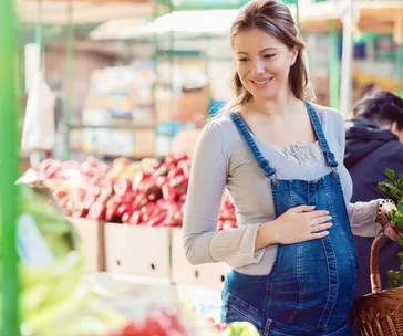 Pregnant woman at a market wearing denim overalls, smiling and holding a basket, surrounded by fresh produce.