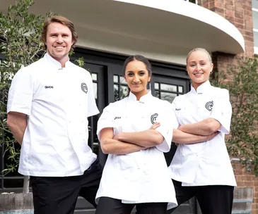 Three chefs in white uniforms standing outside a building, smiling and facing the camera.