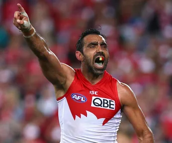 AFL player in red and white jersey, mid-action with arm raised and mouthguard visible, surrounded by blurred crowd.