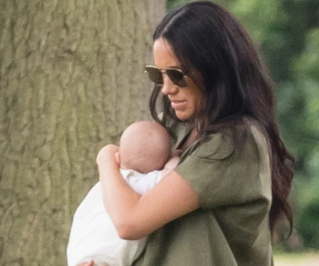 A woman in sunglasses holds a baby outdoors, standing near a tree.