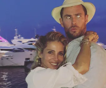 Couple embraces near yachts at twilight; one wears a hat, the other smiles, with boats and pink-lit backdrop in view.