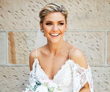 A smiling bride in a lace wedding dress holds a bouquet, standing against a textured stone wall.