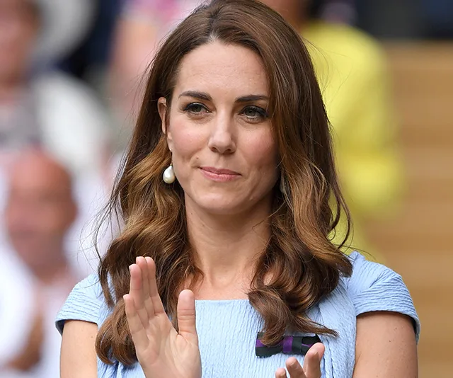A woman with long brown hair clapping, wearing a light blue dress and pearl earrings.