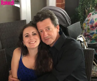 A young woman in a blue dress and an older man in a dark shirt sit closely together, smiling for the camera.