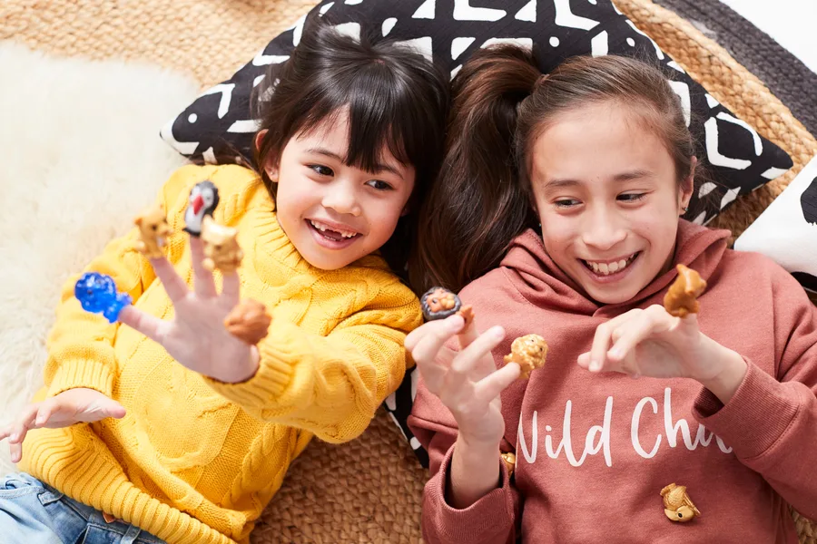 Two children smiling and playing with small collectible figures on their fingers, lying on a cozy surface.