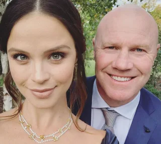A woman in a necklace and a man in a suit smile closely outdoors, with trees in the background.