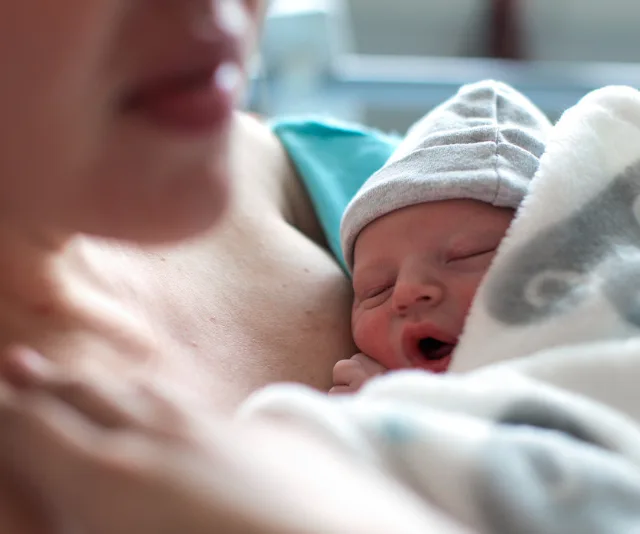 A newborn baby, wearing a hat, sleeps contently on the mother's bare chest, wrapped in a soft blanket.