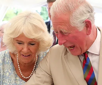 Two people are smiling and looking down at an event, under a tent with trees in the background.