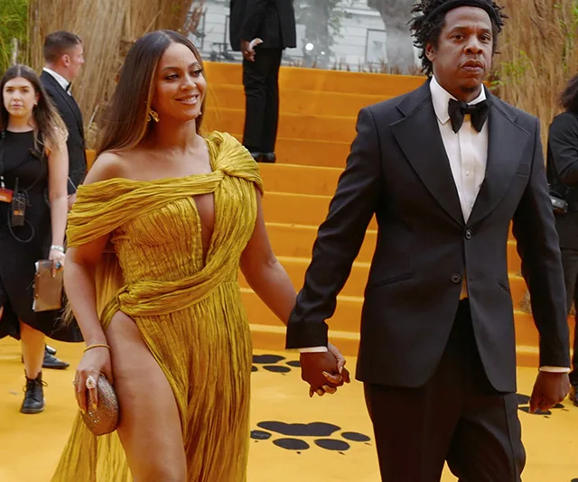 Couple holding hands on an orange carpet; woman in yellow dress, man in black tuxedo.