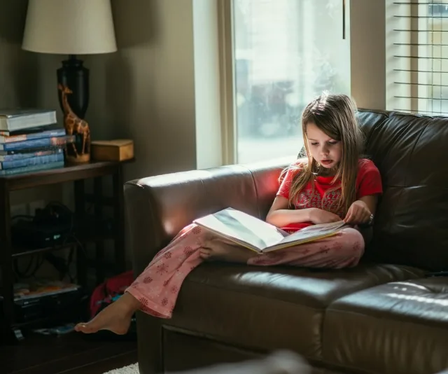 Child in pajamas sitting on a couch, reading a large book with sunlight streaming through a nearby window.