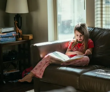 Child in pajamas sitting on a couch, reading a large book with sunlight streaming through a nearby window.