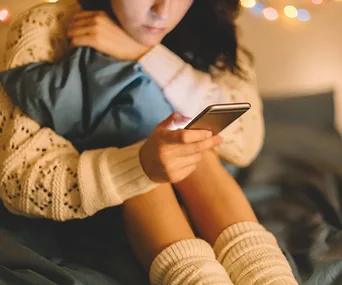 A person in a sweater and leg warmers looks at a smartphone screen while sitting on a bed with cozy lights in the background.